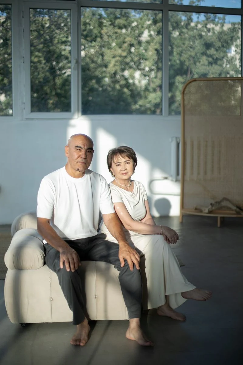 Mutuelle Sante Senior : An elderly couple sitting together on a sofa indoors, illuminated by soft natural light.