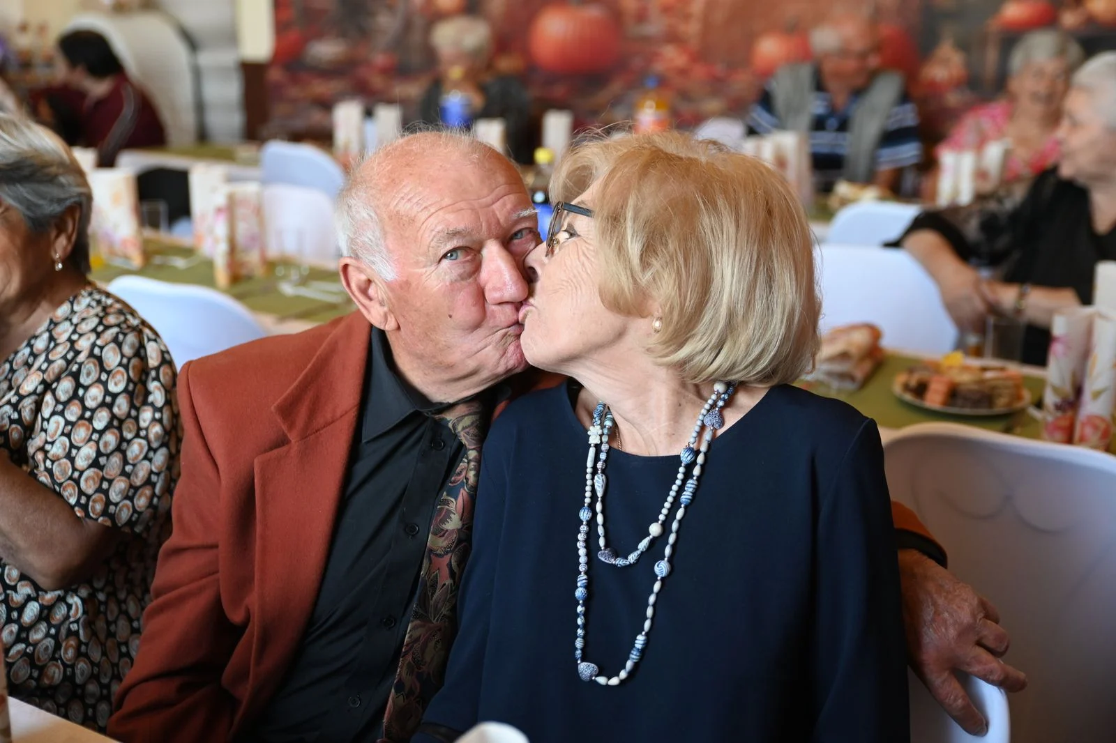 Mutuelle Sante Senior : An elderly couple lovingly shares a kiss at a friendly social event indoors.
