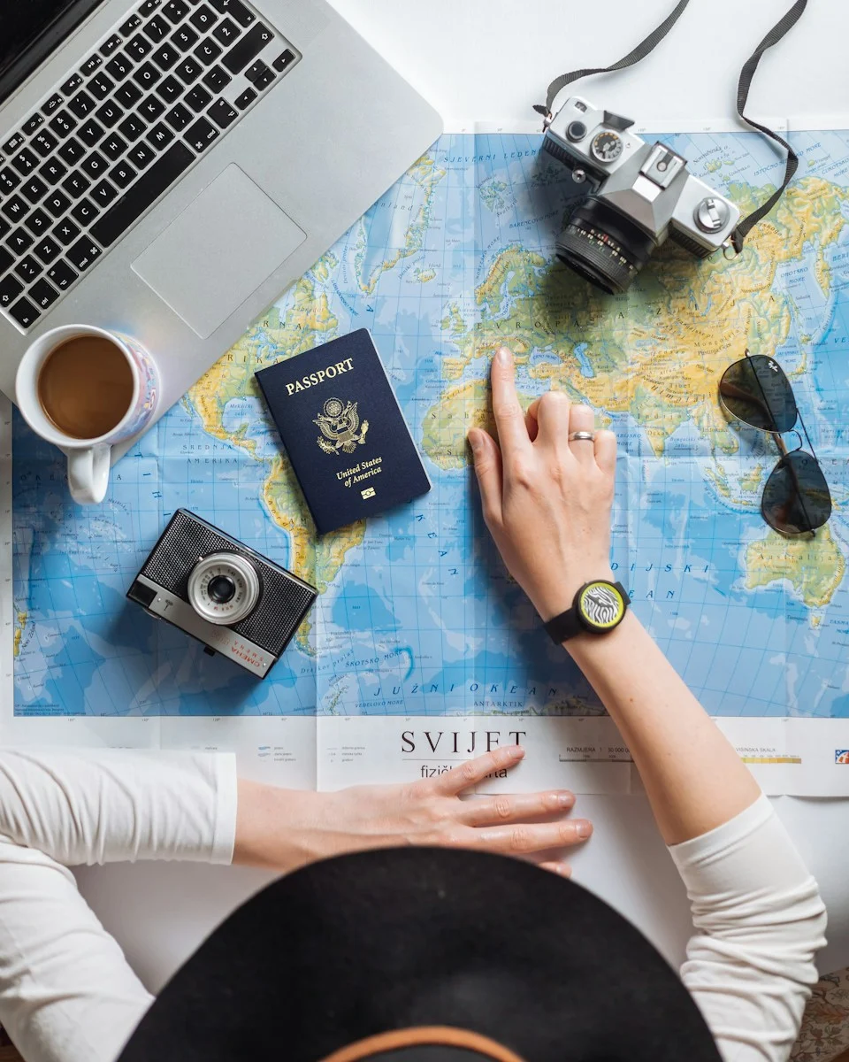Expatrie : A person pointing at a world map with a passport, cameras, and laptop